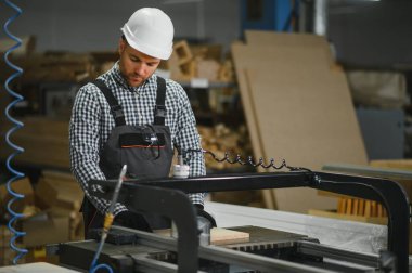 Marangoz güvenlik kaskı takıyor ve işçi tulumu giyiyor. Mobilya fabrikasında bir makine çalıştırıyor. Kanepe üretimi için tahta kesiyor.