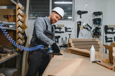 Marangoz güvenlik kaskı takıyor ve tulum giyiyor. Pnömatik çivi tabancasıyla bir atölyede ahşap mobilya parçaları birleştiriyor.