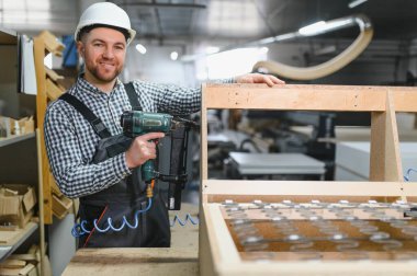 Gülümseyen marangoz elinde pnömatik çivi tabancası tutarken mobilya atölyesinde kanepe çerçevesi kuruyor, marangozluk yeteneklerini sergiliyor.