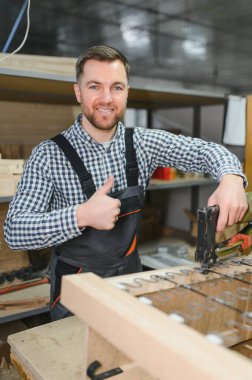 Gülümseyen marangoz ahşap kanepe çerçevesini monte ediyor. Pnömatik zımba tabancası kullanıyor. Mobilya fabrikasında baş parmak işareti yapıyor.