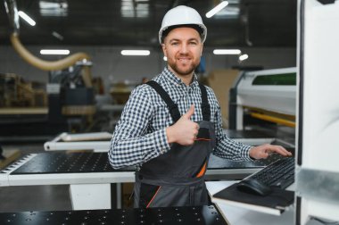 Marangoz kask ve tulum giyer, bir CNC makinesi çalıştırır ve bir mobilya fabrikasında baş parmağını kaldırır.