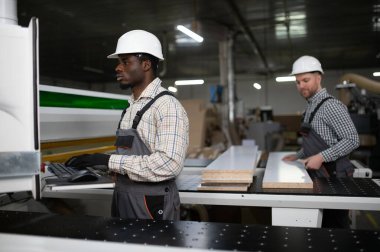 İki fabrika işçisi bir CNC makinesi çalıştırıyor, mobilya parçaları üretiyorlar, takım çalışması ve modern üretim süreçlerini sergiliyorlar.