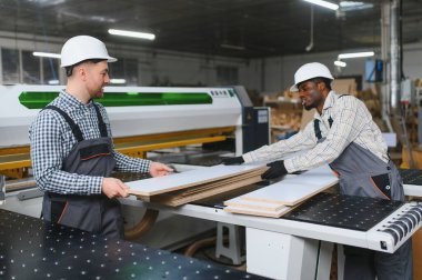 İki farklı fabrika işçisi, modern bir mobilya fabrikasında kanepe üretiminde işbirliği yaparak, bir makinenin yanında işlenmiş ahşap tahtaları dikkatle tutuyorlar.