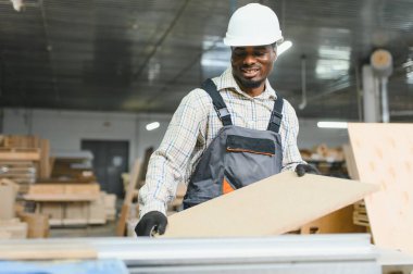 Gülümseyen Afro-Amerikan marangoz bir mobilya fabrikasında çalışırken ahşap bir panel tutuyor, marangozluk ve kendini adamışlık sergiliyor.
