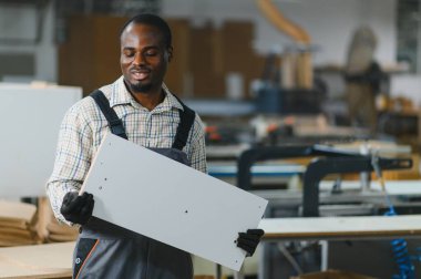 Telaşlı bir mobilya fabrikasında çalışırken ahşap bir tahtayı tutan gülümseyen marangoz, iş başında marifetli zanaatkarlık sergiliyor.