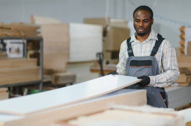 Odaklanmış Afro-Amerikan marangoz ahşap tahtalarla çalışıyor, bir mobilya fabrikasında üretim sürecini yönetiyor.