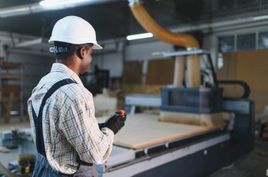 Marangoz, sıkı bir şapka ve eldiven takıyor. Mobilya fabrikasında ustalıkla cnc makinesi kullanıyor. Hassas ahşap ürünler üretiyor.