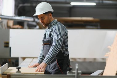 Marangoz güvenlik kaskı takıyor ve iş tulumları ile mobilya fabrikasındaki modern Cnc makinesine ahşap paneli dikkatlice yerleştiriyor, kesin kesim ve şekillendirmeye hazırlanıyor.