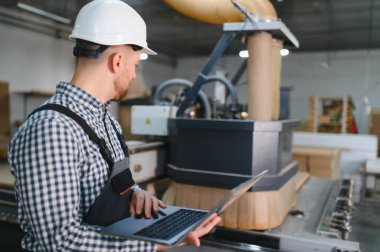 Modern bir mobilya fabrikasında dizüstü bilgisayar kullanarak kanepe üretimi ve ahşap işçiliği üzerine çalışan yetenekli bir işçi.