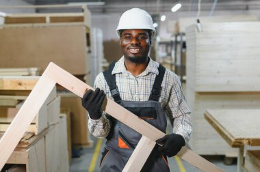 Güvenlik kaskı takan gülümseyen marangoz, bir mobilya fabrikasında çalışırken ahşap bir kalas tutuyor ve marangozluk yeteneği sergiliyor.