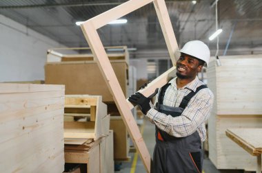 Gülümseyen marangoz mobilya fabrikasında ahşap çerçeve taşıyor, el işi ve endüstriyel üretim sergiliyor.