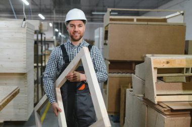 Gülümseyen marangoz ahşap bir çerçeve tutuyor, çalışkan bir mobilya fabrikasında özenle çalışıyor, uzmanlık ve beceri sergiliyor.