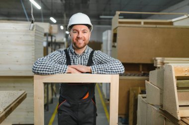 Marangoz, mobilya fabrikasında güler yüzlü ve ahşap çerçeveye yaslanır, el işi ve kalite kontrolü sergiler.