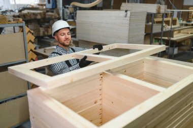 Marangoz güvenlik kaskı ve eldiven takıyor mobilya parçalarının üzerine ahşap çerçeve yerleştiriyor.
