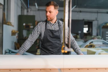 Bir mobilya atölyesinde büyük bir rulo beyaz kumaş kesmek için elektrikli testereyle önlük giyen marangoz.