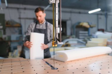 Marangoz, yoğun bir mobilya fabrikasında elektrikli testereyle koltuklar için köpük kesiyor, maharetli işçilik ve üretim hassasiyeti sergiliyor.