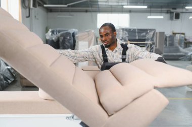 Mobilyacı, hareketli bir fabrikada şık bir kanepe kuruyor, marifetli işçilik sergiliyor ve mobilya üretiminde kaliteye kendini adamış.