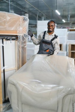 Gülümseyen Afrikalı Amerikalı fabrika işçisi mobilya üretim hattı, kalite kontrol ve endüstriyel üretim konseptine sarılı kanepeyi gösteriyor.