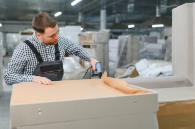 Marangoz, mobilya fabrikasındaki ahşap kanepe çerçevesine koruyucu kağıt takmak için zımba tabancası kullanıyor.