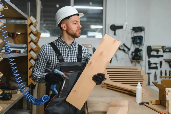 Marangoz ahşap kalası inceliyor, pnömatik çivi tabancası tutuyor, mobilya fabrikasında, aletler ve malzemelerle çevrili.