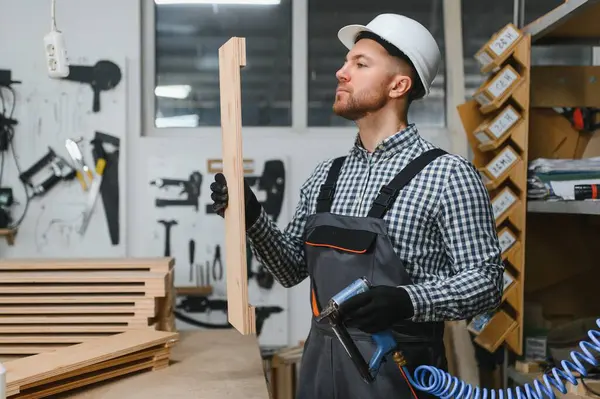 Marangoz koruyucu iş giysisi giyip ahşap bir kalası incelerken bir mobilya fabrikasında pnömatik çivi tabancası tutuyor.