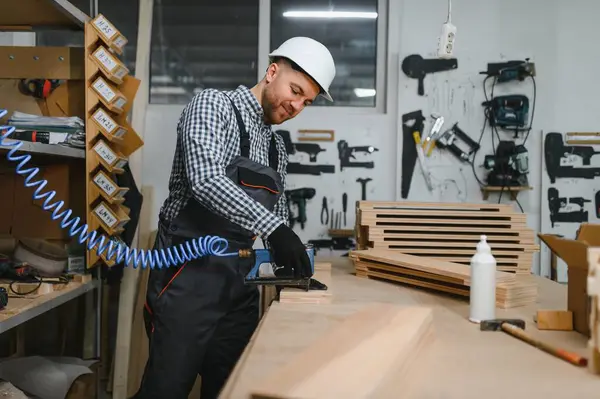Marangoz güvenlik kaskı takıyor ve tulum giyiyor. Pnömatik çivi tabancasıyla bir atölyede ahşap mobilya parçaları birleştiriyor.