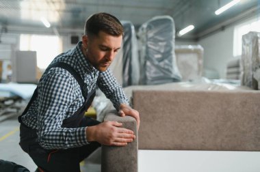 Çalışanlar, malzeme ve makinelerle çevrili bir mobilya fabrikasında yeni üretilen kanepeyi dikkatlice inceleyerek kalite ve hassasiyeti sağlıyorlar.