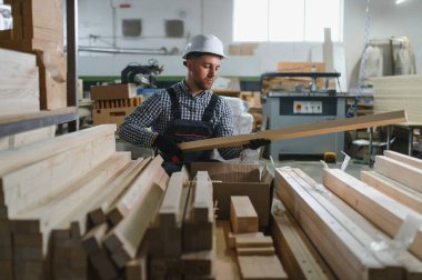 Marangoz güvenlik kaskı takıyor. Bir mobilya fabrikasında ahşap kalasları inceliyor. Etrafı çeşitli tahta parçaları ve makinelerle çevrili.