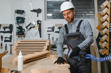 Marangoz, pnömatik çivi tabancası kullanıyor. Ahşap mobilyaları ustalıkla, malzeme ve aletlerle dolu, hareketli bir atölyede monte ediyor.