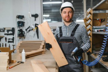 Ahşap bir tahta ve çivi tabancası tutan marangoz, yoğun bir atölyede mobilya montajına hazırlanıyor.