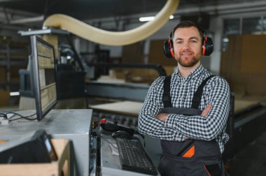 Ses geçirmez kulaklık takan ve mobilya atölyesinde kolları çapraz duran gülümseyen bir marangoz portresi.