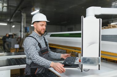 Bir mobilya fabrikasında güvenlik kaskı takan marangoz bilgisayar kullanarak ahşap işlemeyi kontrol ediyor.