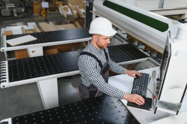 Mobilya fabrikasında güvenlik kaskı takan marangoz, işlemi bilgisayar aracılığıyla kontrol ediyor.