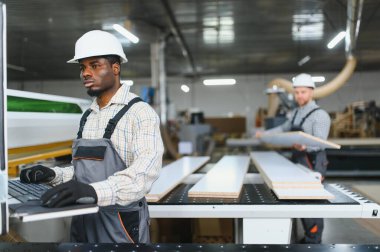 İki marangoz, bir mobilya fabrikasında ahşap tahtalar kesmek için bir makineyle çalışıyor.