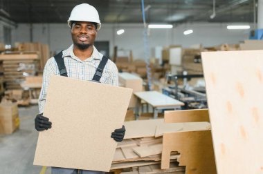 Afrika kökenli Amerikalı işçiler işlek bir mobilya fabrikasında ahşap bir panel tutuyor. Etrafı malzeme ve makinelerle çevrili. Üretim süreçlerini gösteriyor.