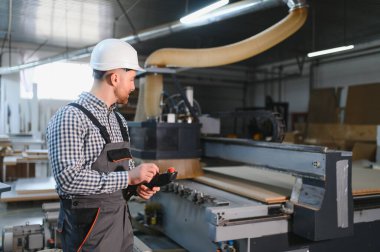 Marangoz güvenlik kaskı takıyor ve bir mobilya fabrikasında CNC makinesini çalıştırıyor, süreci uzaktan kumandayla kontrol ediyor.