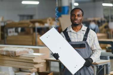 Afro-Amerikan marangoz bir mobilya fabrikasında beyaz ahşap bir tahta tutuyor.