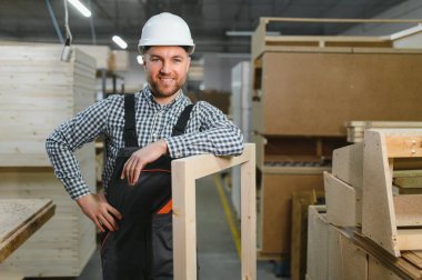 Kendine güvenen marangoz ahşap bir çerçeveye gururla yaslanır, hareketli bir mobilya fabrikasında işçilik sergiler.