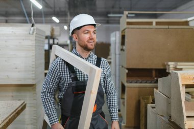 Gülümseyen marangoz güvenlik kaskı takıyor ve işlek mobilya fabrikasında çalışırken ahşap bir çerçeve taşıyor.