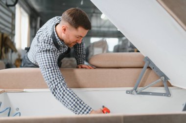 Marangoz bir mobilya fabrikasında kanepe monte ediyor, ahşap ve döşemelerle çalışırken kaliteli işçiliği vurguluyor.