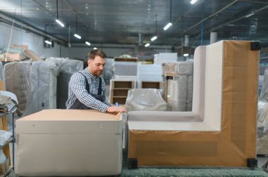 Marangoz mobilya fabrikasında çalışıyor, kanepe parçaları monte ediyor koruyucu bant ve karton kullanarak, bitmiş ürünlerle çevrili