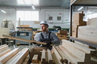 Marangoz koruyucu iş kıyafeti giyiyor, mobilya fabrikasında ahşap bir kalası inceliyor, üretim sürecinde kalite sağlıyor.