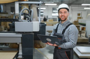 Marangoz bir dizüstü bilgisayarla bir cnc tahta kesme makinesini kontrol ediyor, bir mobilya fabrikasında çalışıyor.