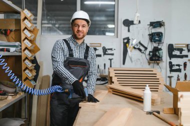 Marangoz güvenlik kaskı takıyor. Pnömatik çivi tabancası kullanıyor. Atölyede ahşap mobilya parçaları birleştiriyor.