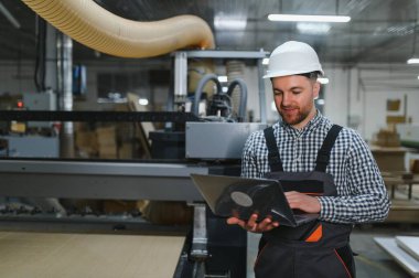 Marangoz, mobilya fabrikasında çalışırken bir dizüstü bilgisayarla ahşap yönlendirici programlıyor. Teknolojiyi geleneksel işçilikle harmanlıyor.