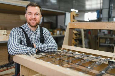 Marangoz, mobilya fabrikasında yayları olan ahşap kanepeye yaslanırken kameraya gülümsüyor.