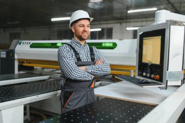 Marangoz güvenlik kaskı takıyor ve iş tulumu giyiyor. Mobilya fabrikasında çapraz kollarla gülümsüyor.