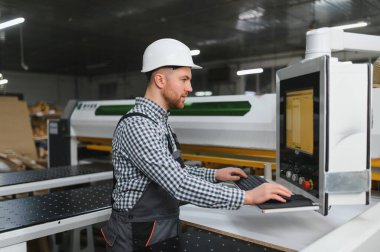 Mobilya fabrikasında güvenlik kaskı takan marangoz otomatik üretim sürecini kontrol ediyor.