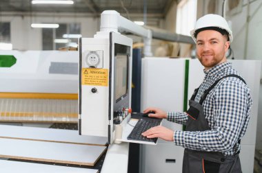 Marangoz, ahşap fabrikasındaki mobilya üretimi için bir Cnc makinesinin kontrol panelini kullanarak kasketini takıyor.