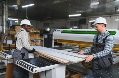 İki fabrika işçisi, ahşap kalasları dikkatle tutuyor. Onları modern bir mobilya fabrikasında, cnc makineli kanepe üretimi için hazırlıyorlar.